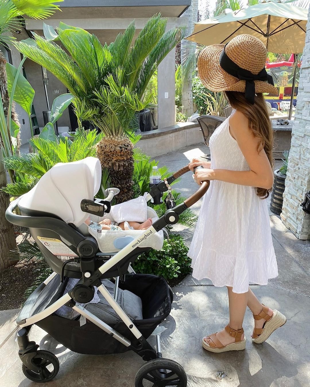 A woman in a white dress and straw hat pushes a black and white stroller with a baby inside in an outdoor patio setting with plants.