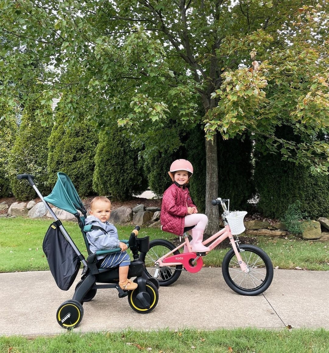 A toddler sits in a black stroller with a teal canopy outdoors on a paved path, while an older child rides a pink bicycle beside it. Trees and shrubs are visible in the background.