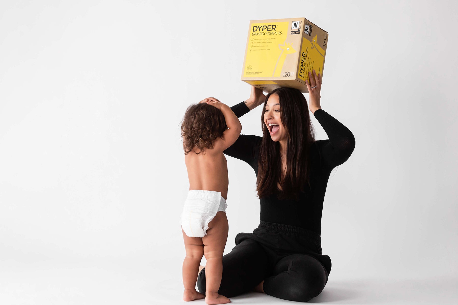 Mom holding a box of disposable diapers over her head next to her baby who is wearing a disposable diaper