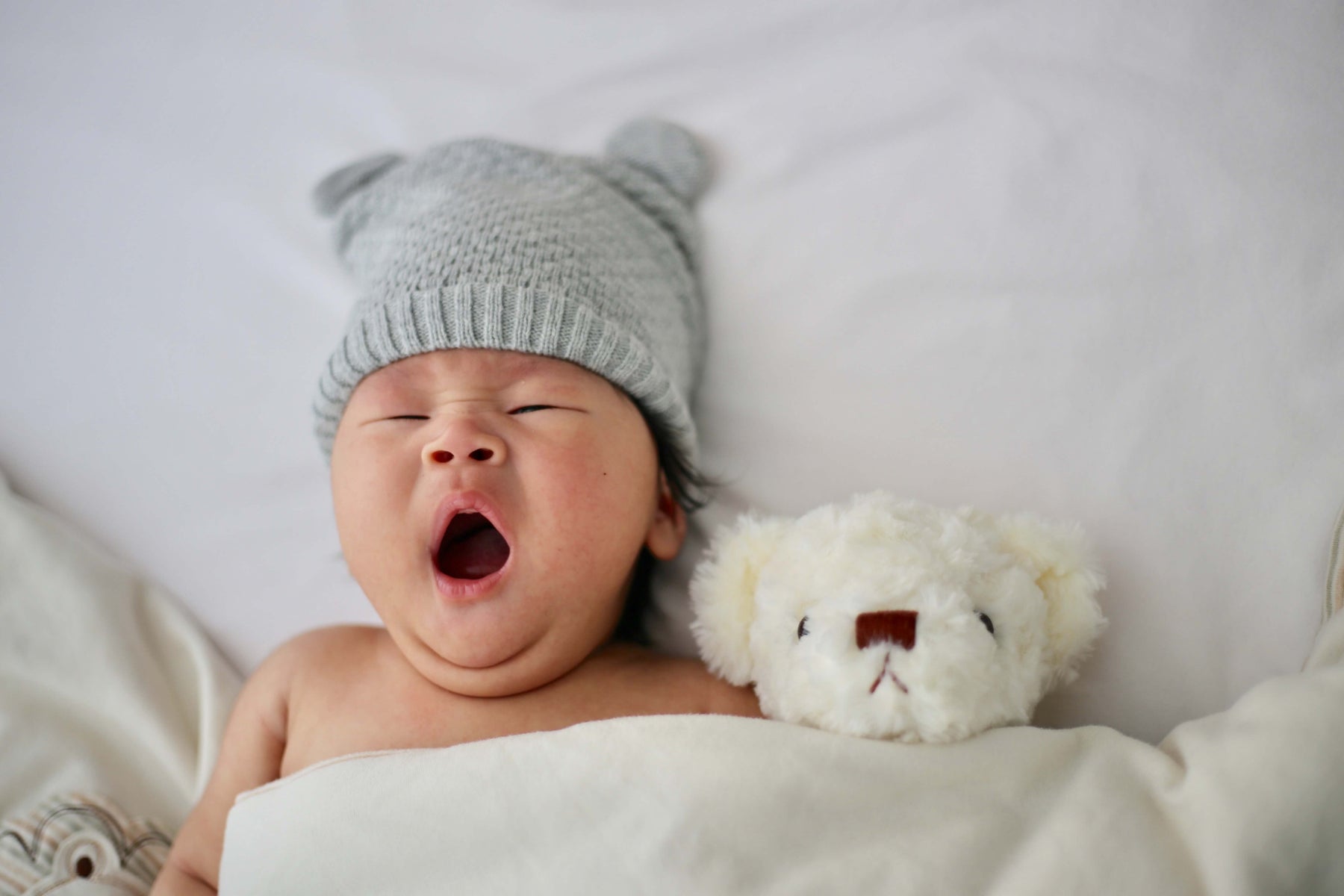 baby laying in bed yawning with a beanie on and a stuffed animal