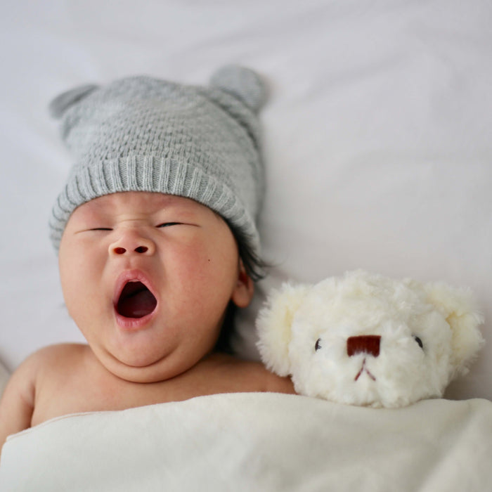 baby laying in bed yawning with a beanie on and a stuffed animal