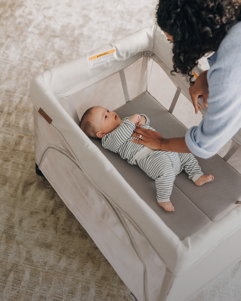 A baby in a striped one-piece outfit lies on a padded changing surface inside a light beige fabric playard while an adult in a light blue top leans over the crib in a softly lit indoor setting.