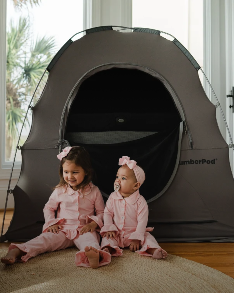 Two young children in matching pink outfits and bows sit in front of a gray SlumberPod tent on a rug in a bright indoor room.