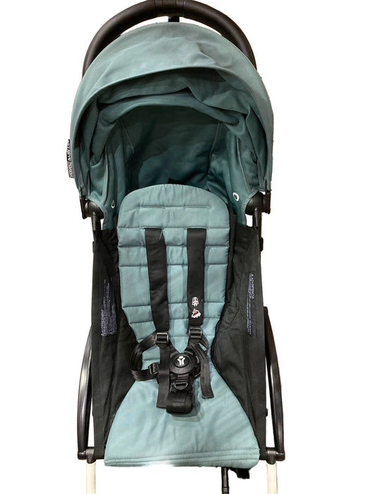 Teal baby stroller with black straps on a gray floor with cardboard boxes in the background