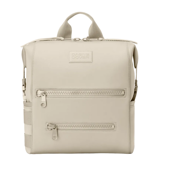 A light beige Dagne Dover diaper backpack with dual top handles, two front zippered pockets, and side pockets, shown against a plain white background.