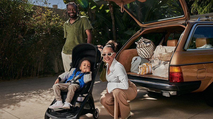 A family scene with a man standing behind a stroller, a woman crouching beside it, and an open station wagon loaded with bags in a sunny outdoor setting.