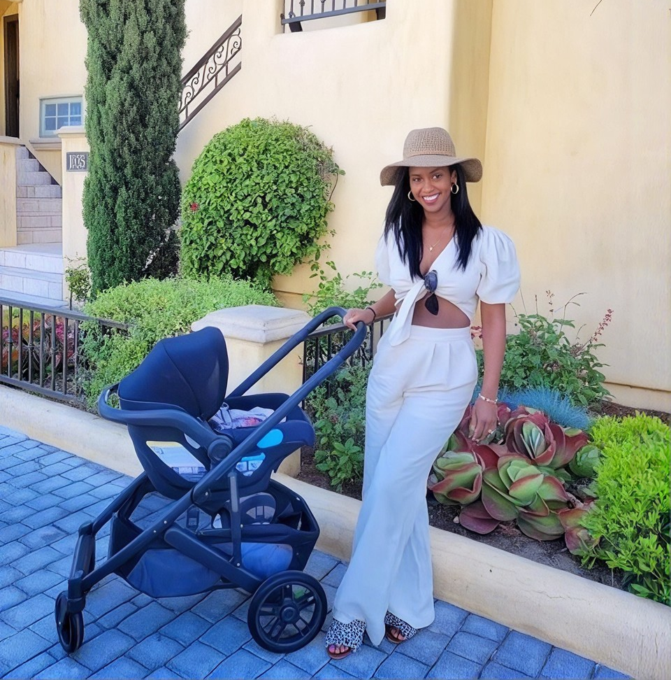 A woman wearing a white two-piece crop top and wide-leg pants stands next to a navy blue stroller in front of a beige stucco building with green plants.