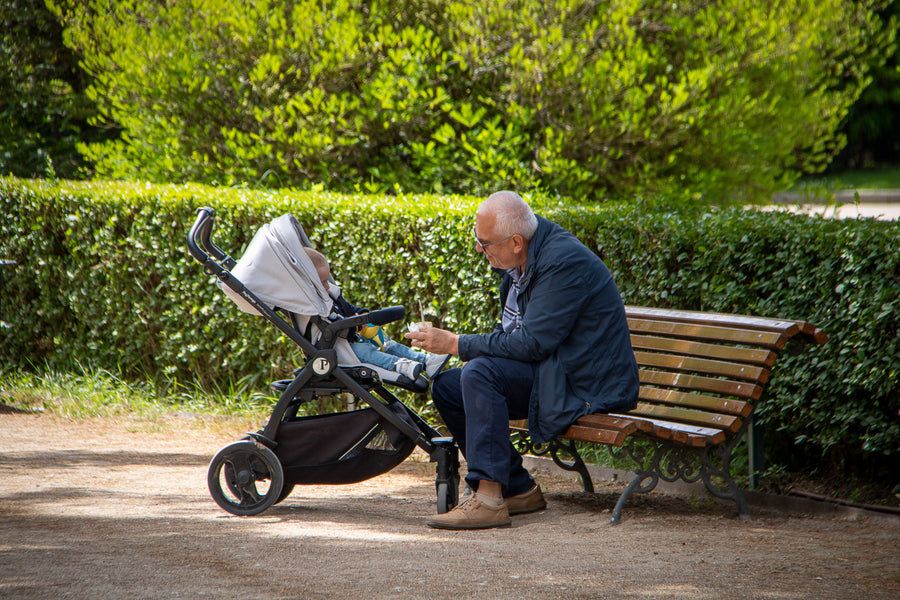 An elderly man sits on a wooden park bench next to a stroller, interacting with a child inside the stroller in a sunny, green park setting.